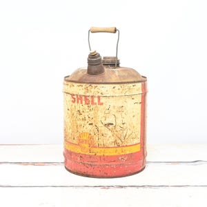 Puede incluir: Bidón de gasolina vintage de metal oxidado Shell con asa de madera. El bidón es amarillo con detalles rojos y negros y la palabra "Shell" impresa en el lateral.