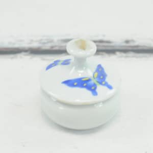 Puede incluir: Un pequeño tarro de cerámica blanca con una tapa que presenta un diseño de mariposa azul. El tarro tiene una forma redonda y un acabado liso y brillante.