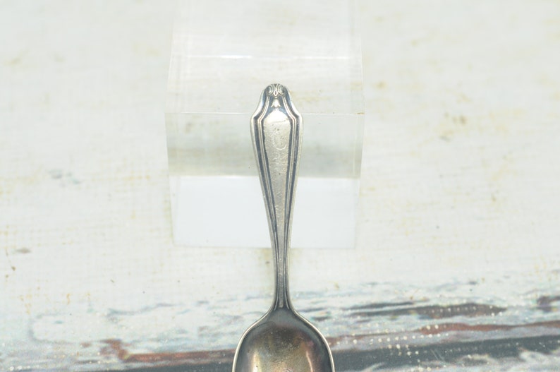 May include: A silver teaspoon with a decorative handle and a rounded bowl. The handle has a raised design and the bowl is slightly dented.