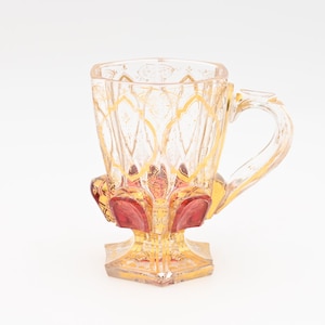 Puede incluir: Una taza de vidrio transparente con un diseño dorado y rojo. La taza tiene un asa y una base ancha.