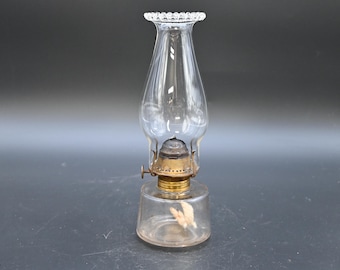 May include: A clear glass oil lamp with a decorative, scalloped top. The lamp has a brass base and a clear glass chimney. The lamp is designed to hold oil and provide light. The lamp is a vintage style.