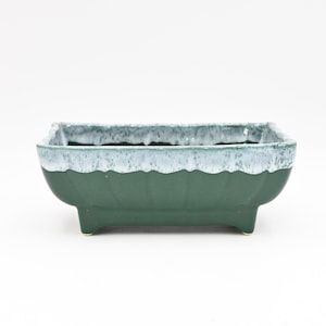 May include: A square green ceramic planter with a white glaze around the top rim. The planter has four small feet and is designed for indoor plants.