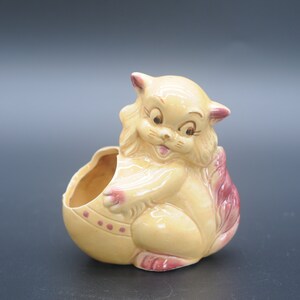 May include: A ceramic planter shaped like a cat with a pink tail. The cat is yellow with brown painted features and is holding an egg-shaped planter.