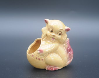 May include: A ceramic planter shaped like a cat with a pink tail. The cat is yellow with brown painted features and is holding an egg-shaped planter.