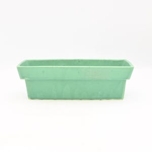 May include: A rectangular, light green ceramic planter. The planter has a simple, geometric design with a slightly flared top and a solid base. The planter is empty and sits on a white surface.