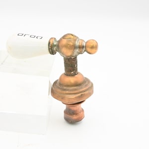 May include: Antique faucet handle with a white ceramic grip and a brass body. The handle has the word "oroa" printed on the ceramic. The faucet is made of brass and copper and is likely from a vintage bathroom fixture.
