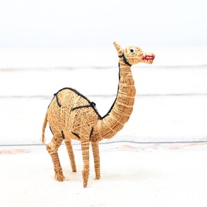 May include: A hand-crafted camel figurine made from natural materials. The camel is brown and has black details. It is standing on a white surface.