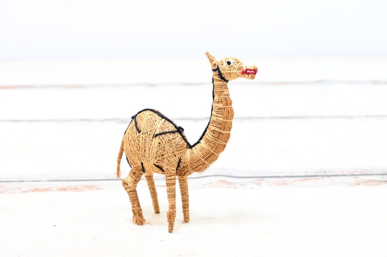 May include: A hand-crafted camel figurine made from natural materials. The camel is brown and has black details. It is standing on a white surface.