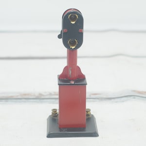 May include: A red and black toy train signal with two gold lights. The signal is on a black base and has a lever on the side.