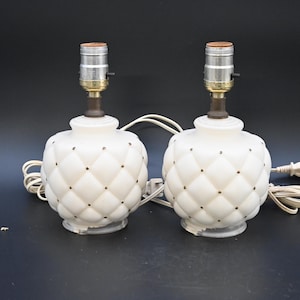 Op de afbeelding: Een paar witte keramische tafellampen met een gestikt ontwerp en gouden accenten. De lampen hebben een eenvoudige, klassieke stijl en zouden een geweldige aanvulling zijn op elk huis.