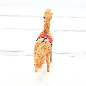 May include: A straw horse figurine with a red and black saddle. The horse is facing away from the camera.