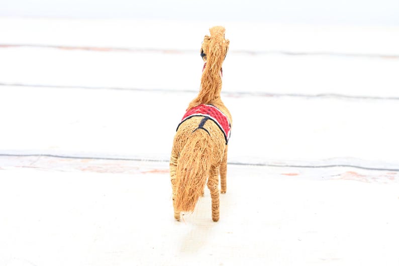 May include: A straw horse figurine with a red and black saddle. The horse is facing away from the camera.