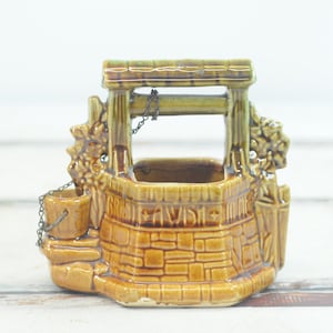 Puede incluir: Un macetero de cerámica con forma de pozo con un esmalte marrón y verde. El pozo tiene un cubo y una cadena, y las palabras "Great Falls, Va." están escritas en la parte delantera.