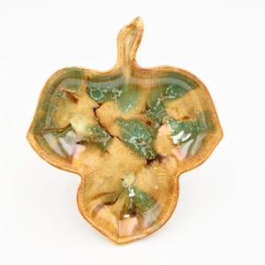 Könnte beinhalten: Eine blattförmige Dekoschale mit natürlicher Holzmaserung. Die Schale hat eine hellbraune Basis mit grünen und weißen Akzenten und ein glänzendes Finish. Die Schale ist wahrscheinlich aus Keramik oder Harz.