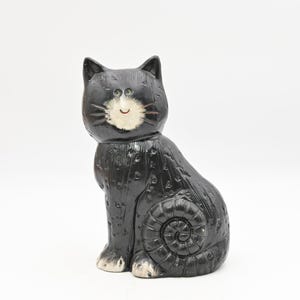 Può includere: Statuetta di gatto in ceramica nera con muso e zampe bianche. Il gatto è seduto in posizione eretta con una superficie strutturata e un motivo a spirale sul lato. Il gatto ha un'espressione sorridente.