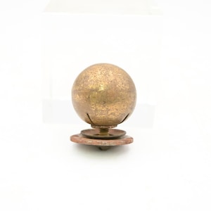 May include: A vintage brass sphere-shaped object with a textured surface, mounted on a circular base. The sphere has a warm, golden hue, and the base appears to be made of a reddish-brown material. The object is likely a decorative item.