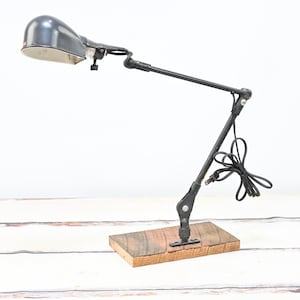 May include: A vintage black desk lamp with an adjustable arm and a wooden base. The lamp has a grey lampshade and a black cord. The lamp is designed to be adjustable and is suitable for a desk or table.