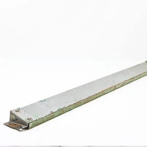 May include: A long, narrow, rectangular metal strip with a weathered grey finish and visible green paint residue along the edges. The object features a flat surface, slightly angled sides, and small metal mounting brackets at both ends with rivets.