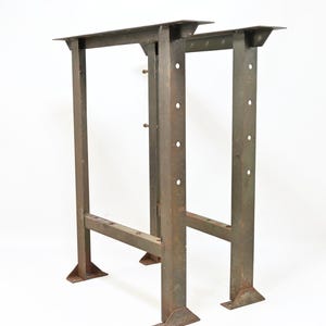 May include: A pair of tall, industrial-style metal table legs featuring a weathered, dark brown finish. Each leg assembly consists of two vertical posts connected by a horizontal crossbar, with flat mounting plates at the top and wide, triangular feet at the base. The vertical posts are punctuated with a series of evenly spaced circular holes along their length.
