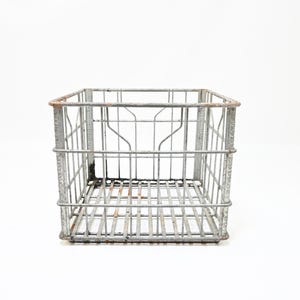 Puede incluir: Una caja de almacenamiento cuadrada de parte superior abierta construida con alambre de metal gris. La estructura presenta un patrón de rejilla con barras verticales y horizontales, mostrando signos visibles de oxidación y óxido en los bordes y la base.