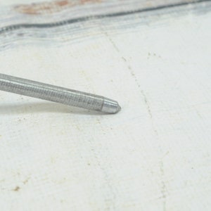 May include: A close-up of a silver metal awl with a pointed tip. The awl is lying on a white surface.