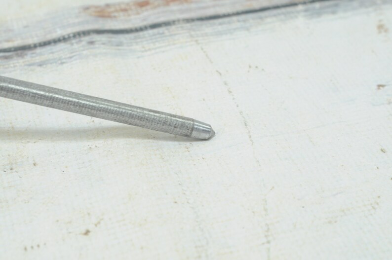 May include: A close-up of a silver metal awl with a pointed tip. The awl is lying on a white surface.