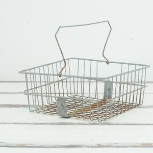 May include: A silver wire basket with a handle. The basket is empty and sitting on a white wooden surface.