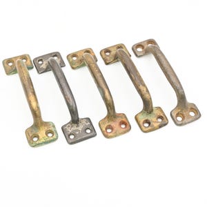 May include: Five antique metal handles with a weathered appearance. The handles are a mix of brass and dark metal, showing signs of oxidation and wear. Each handle has a rectangular base with two screw holes and a curved grip.