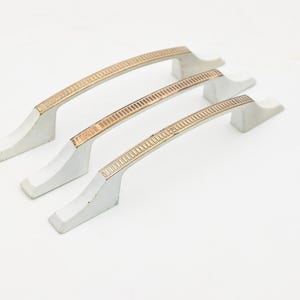 May include: Three cabinet handles with a curved design. The handles are silver with a gold-coloured, rectangular patterned accent. The handles are designed for furniture and have a modern aesthetic.