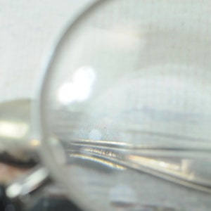 May include: A close-up of a silver spoon under a magnifying glass. The spoon has a detailed design and a rounded handle.