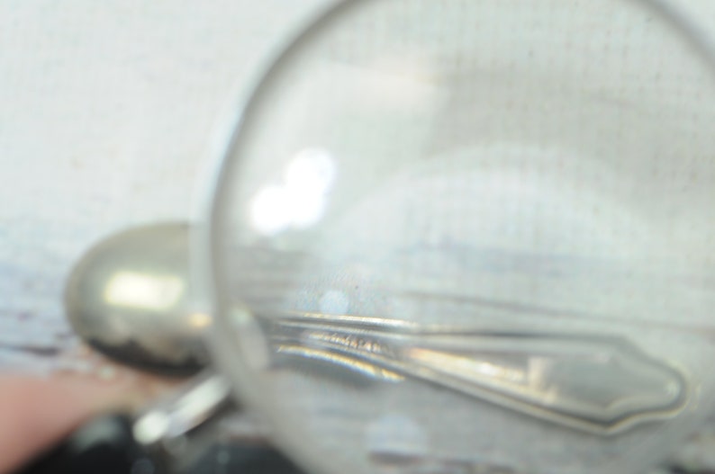 May include: A close-up of a silver spoon under a magnifying glass. The spoon has a detailed design and a rounded handle.
