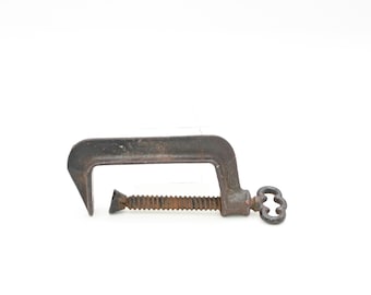 May include: A vintage C-clamp made of dark metal, showing signs of age and use. The clamp features a threaded screw mechanism for adjusting the grip and a decorative handle. The tool is designed for woodworking and construction projects.