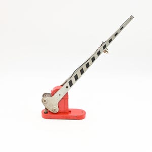 May include: A vintage model railroad crossing gate. The base is red, with a silver arm featuring black and white diagonal stripes. The gate is raised, suggesting an open crossing.