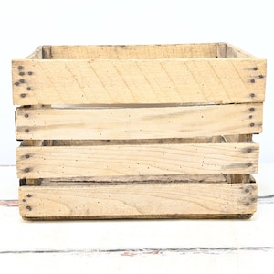 May include: A square, light-colored wooden crate with visible wood grain and metal nails. The crate is constructed from horizontal planks and is empty, with a rustic, vintage aesthetic. The crate is suitable for storage or display.