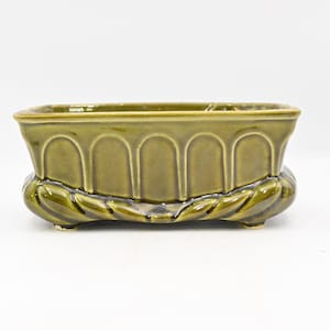 Puede incluir: Una jardinera rectangular de cerámica verde oliva con acabado brillante. La jardinera presenta detalles arqueados en los laterales y una base decorativa con forma de hojas. Ideal para plantas pequeñas.