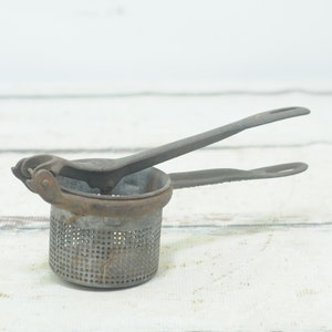 May include: A vintage metal potato masher with a handle and a perforated bowl. The masher is made of metal and has a rustic, aged look.