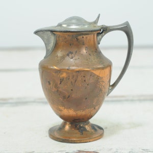 May include: A small copper creamer with a pewter lid and handle. The creamer has a rounded body and a flared base.