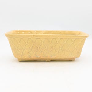 May include: A rectangular planter with a light yellow glazed finish. The exterior features a raised lattice or diamond pattern across all four sides. The vessel has a wide, open top and a slightly tapered base with a small, raised footed edge.