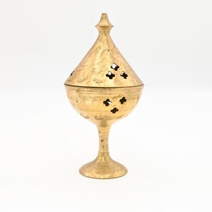 May include: A gold-colored, ornate incense burner with a conical lid and a pedestal base. The lid and body feature floral and geometric cutouts. The surface is etched with intricate patterns. The burner is made of metal and stands on a white surface.