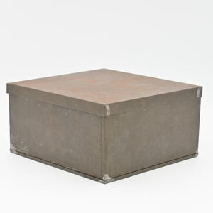 May include: A square, gray metal box with a slightly textured surface. The box is closed and has a simple design.