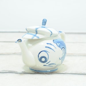May include: A white ceramic teapot with a blue and white patterned design. The teapot has a lid shaped like a hat and a cartoon face with a large eye.