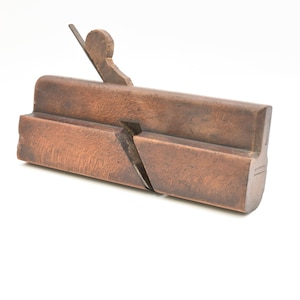 May include: A vintage wooden hand plane with a dark brown finish. The tool features a blade and a handle, and is designed for woodworking. The plane is set against a white background.