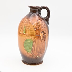 May include: Brown ceramic jug with a handle, featuring a relief of a monk holding a glass and a bottle. The jug has a detailed design of a brick wall, a green barrel, and bottles. The jug is approximately 10 inches tall.