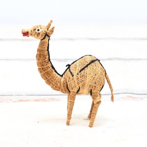 May include: A small, hand-crafted camel figurine made from natural materials. The camel is brown and has a black saddle. It is standing on a white surface.