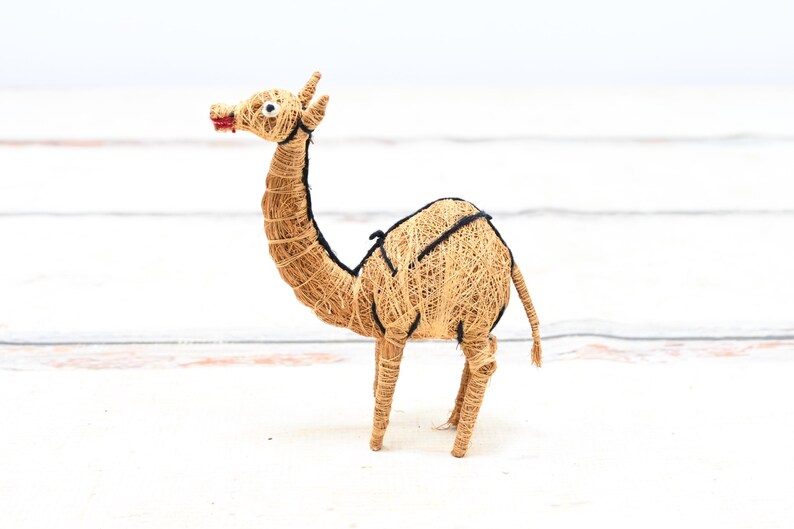 May include: A small, hand-crafted camel figurine made from natural materials. The camel is brown and has a black saddle. It is standing on a white surface.