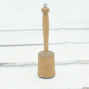 Puede incluir: Un machacador de patatas de madera con una cabeza redonda y un mango largo. El mango tiene un pequeño bucle en la parte superior.