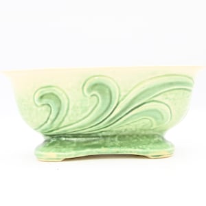 以下が含まれることがあります： 淡い緑色とクリーム色の陶器のボウル。濃い緑色の波模様、台座、フレアリムが特徴です。装飾やサービングに最適です。