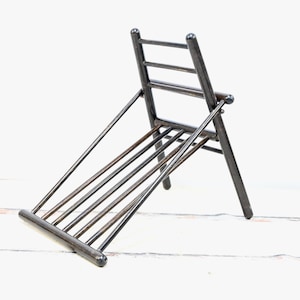 May include: A dark gray, metal folding chair with a ladder-back design. The chair has a slatted seat and is angled, showcasing its minimalist construction. The chair's legs are sturdy, and the overall design is simple and functional.