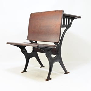 May include: A vintage school desk featuring a dark brown wooden seat and backrest supported by a sturdy black cast iron frame. The design includes a connected writing surface extending from the backrest, supported by decorative metal brackets.