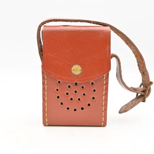 May include: A vintage brown leather case with a flap closure and a strap. The case has a series of small, evenly spaced holes on the front and is secured with a gold-colored button. The strap is also brown and appears worn.